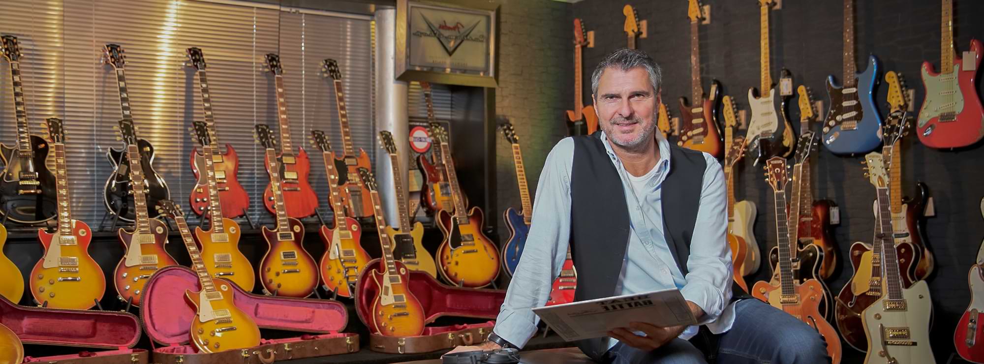 Uli Roth in einem Raum mit über 20 E-Gitarren, hält eine Schallplatte in der Hand
