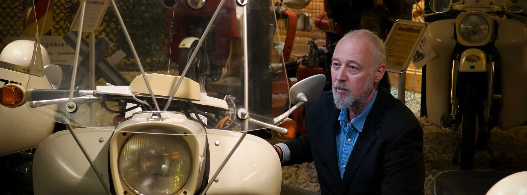 Thomas Müller in einem Motorradmuseum, knieend vor einer Maschine, und bewundert diese