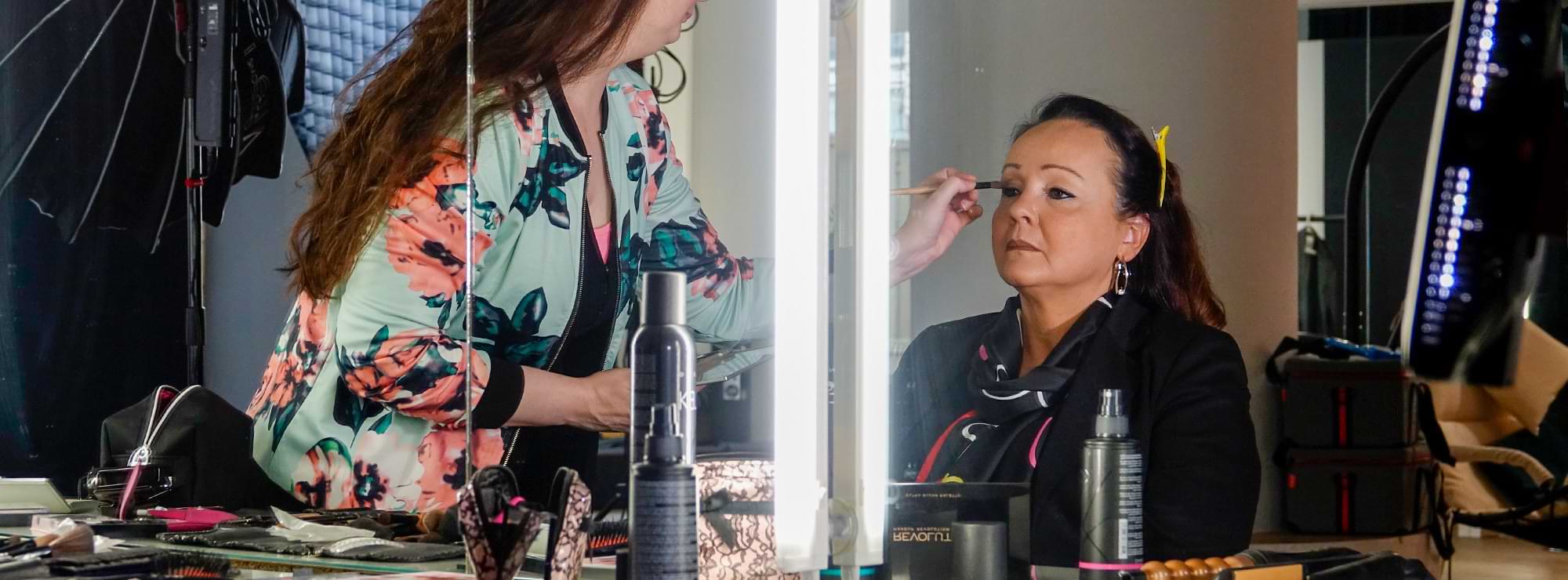 Annette Wenz wird vor einem Spiegel von einer Visagistin geschminkt