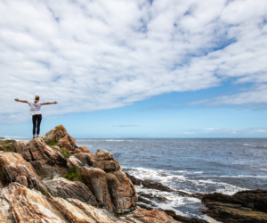 Person mit ausgestreckten Armen steht auf Felsen vor dem Meer