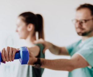 Therapeut weist junge Patientin in Übung mit Gewicht ein. Sie hält das Gewicht am ausgestreckten Arm.