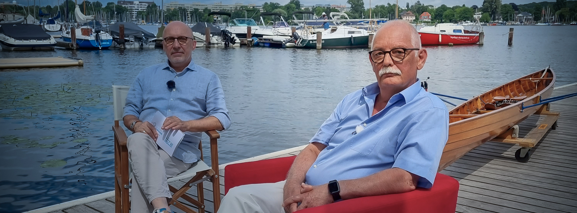 Klaus Eisenbeisz sitzt bei Interview auf dem Roten Sessel an einem Segelboothafen mit dem Moderator, im Hintergrund Schiffe und die Havel