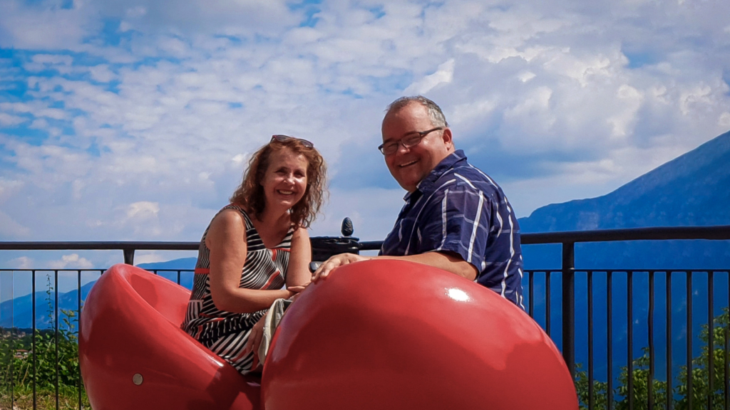 Andreas Cramer und Frau Monika sitzen an einem Aussichtspunkt auf 2 roten Stühlen, im Hintergrund Berge und Himmel