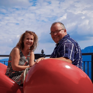 Andreas Cramer und Frau Monika sitzen an einem Aussichtspunkt auf 2 roten Stühlen, im Hintergrund Berge und Himmel