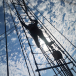 Mast eines Segelboots mit einer Person in den Tauen hängend, Blick Richtung Himmel