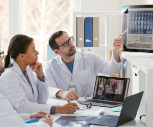 Ein Arzt zeigt auf einen Computerbildschirm und bespricht Röntgenbilder mit seinen Kollegen während andere Ärzte in einer Online-Konferenz zugeschaltet sind