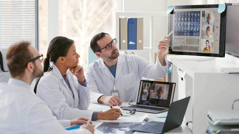 Ein Arzt zeigt auf einen Computerbildschirm und bespricht Röntgenbilder mit seinen Kollegen während andere Ärzte in einer Online-Konferenz zugeschaltet sind